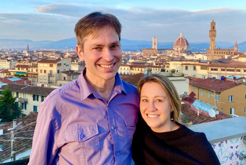 Elon and Yelena Danziger stand on a balcony with a cityscape behind them.