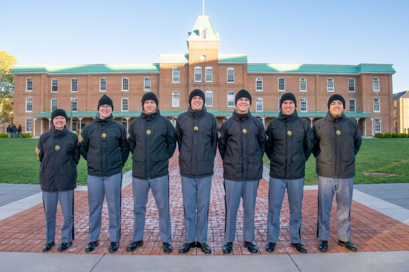The seven cadets are all in uniform with black jackets and black hats on Upper Quad with a historic brick building in the background. 