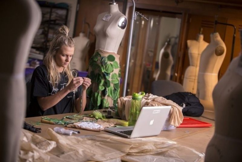 A fashion merchandising and design student sits at a large worktable in a campus maker space, focused on hand-stitching fabric while surrounded by sewing tools, fabric pieces, a laptop, and several dress forms displaying in-progress garment designs.