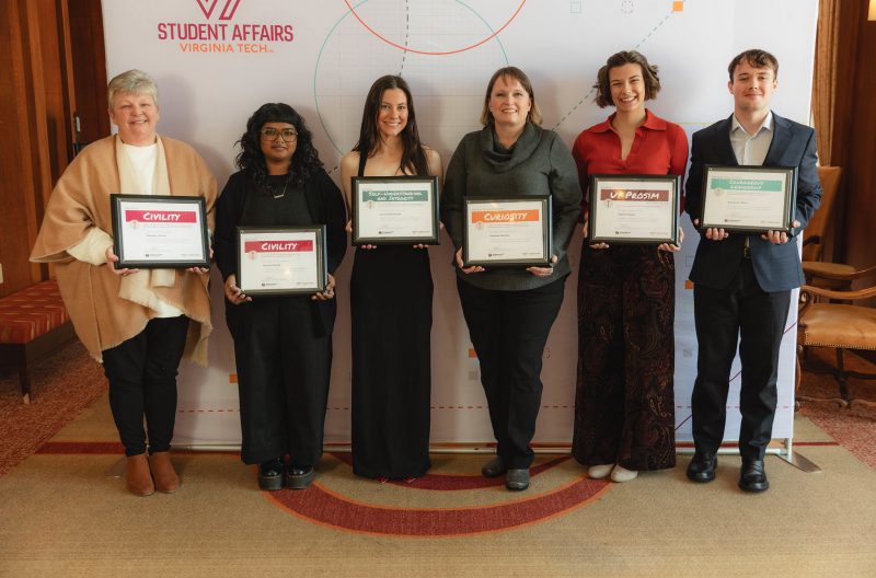 Six smiling people hold variously colored, framed certificates as they stand before a banner labeled Student Affairs.
